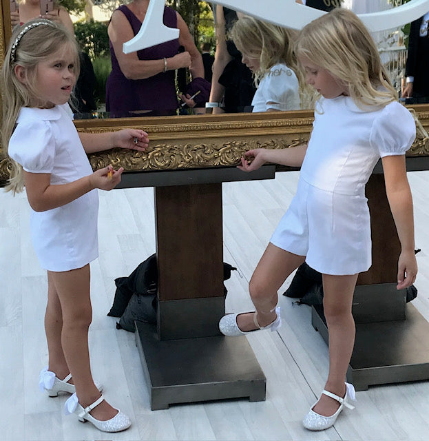 The Melissa romper blends traditional flower girl elegance with playful tap pants. Features puff sleeves, a Peter Pan collar, grosgrain ribbon trim, and easy-to-move shorts. Back pearl button closure with a zipper on the shorts and a hand-tied sash for a polished finish. Handcrafted in Brooklyn from cotton and satin, with optional personalized embroidery. Detachable skirt and custom colors available upon request.