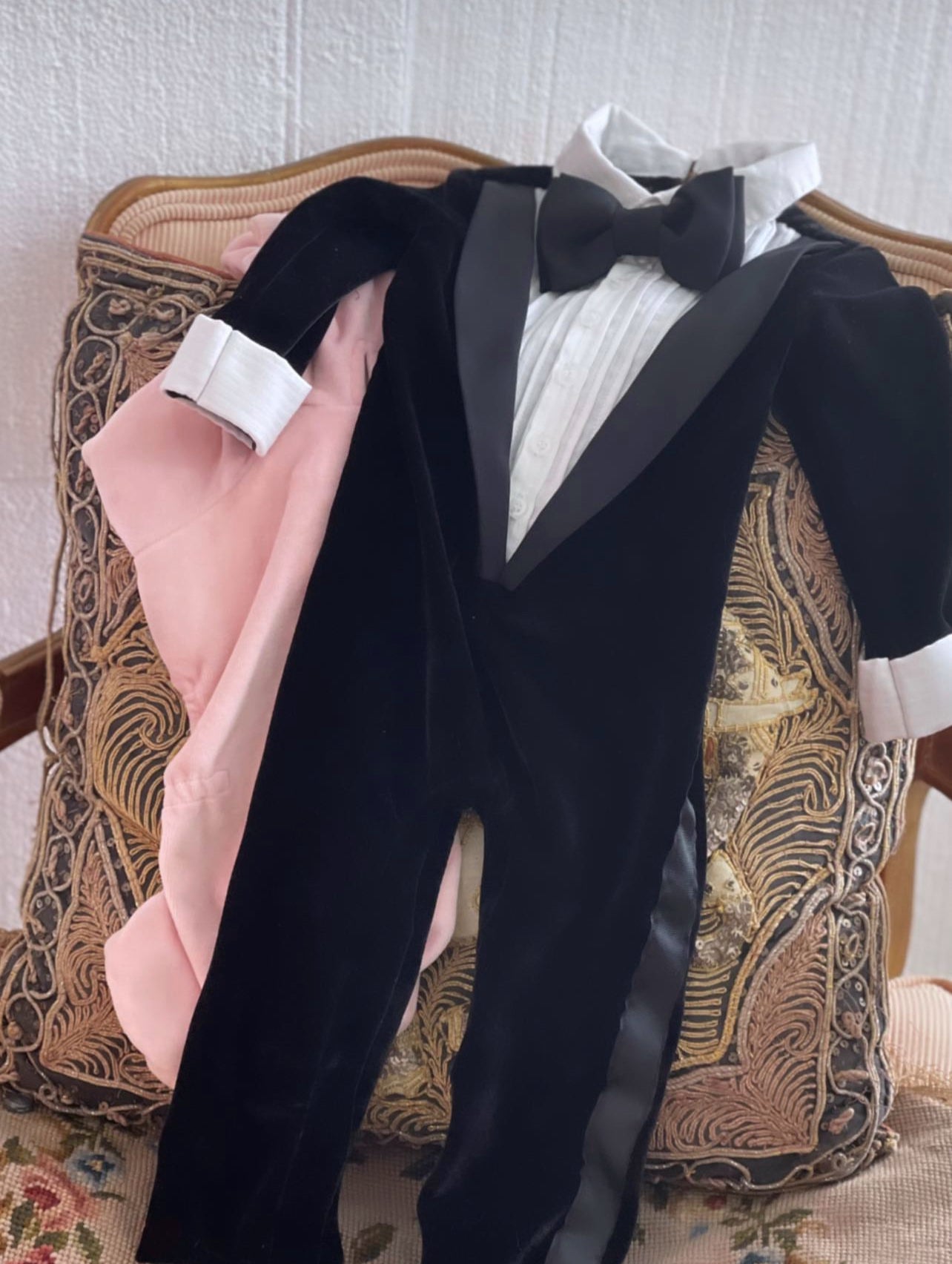 The Tuxedo Onsie, a baby formalwear outfit featuring a black velvet tuxedo design with satin lapels, a crisp white shirt front, and an attached bow tie. Long-sleeved and footless for comfort, perfect for weddings, special occasions, and formal events. Handmade with care.