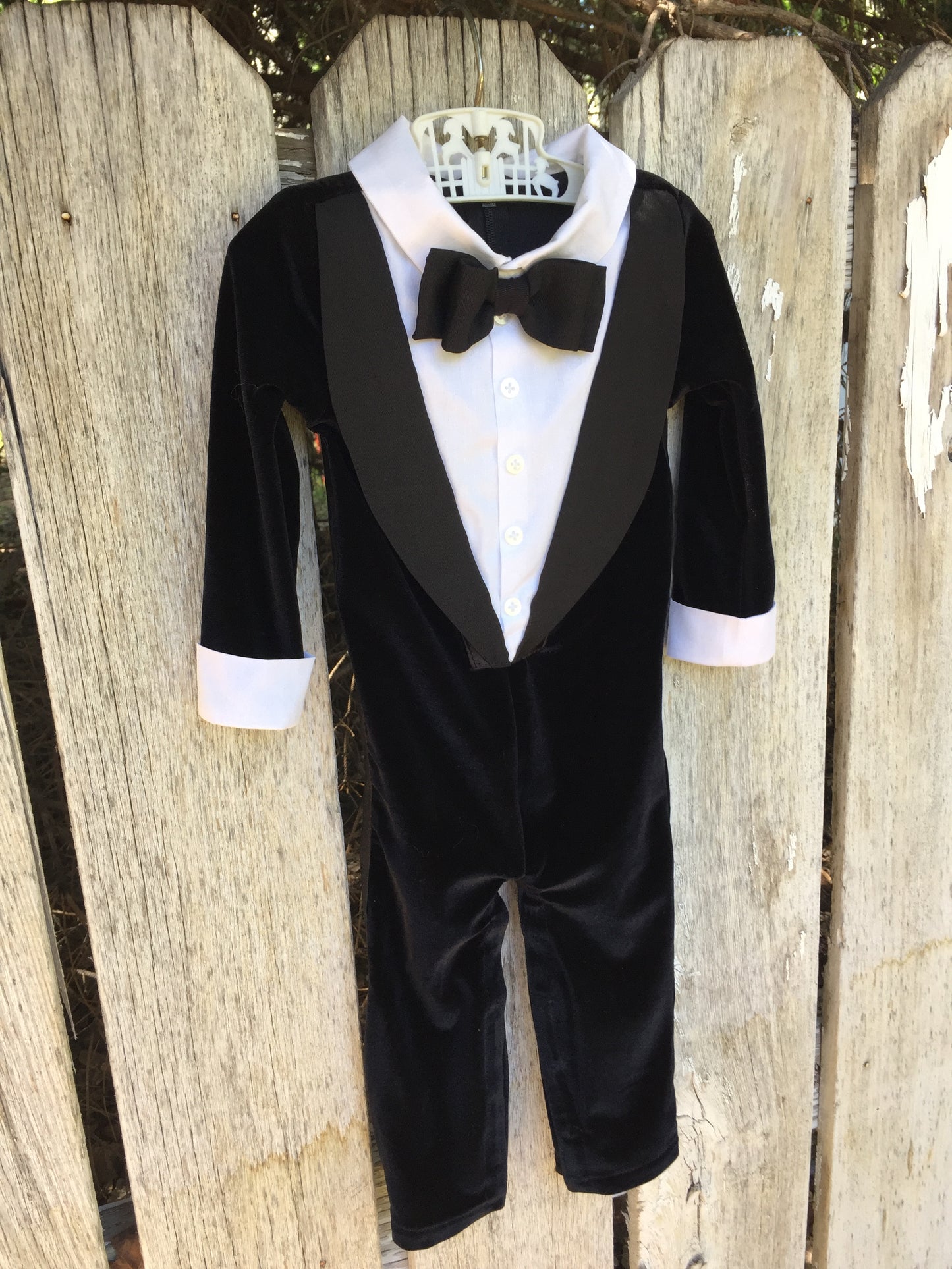 The Tuxedo Onsie, a baby formalwear outfit featuring a black velvet tuxedo design with satin lapels, a crisp white shirt front, and an attached bow tie. Long-sleeved and footless for comfort, perfect for weddings, special occasions, and formal events. Handmade with care.