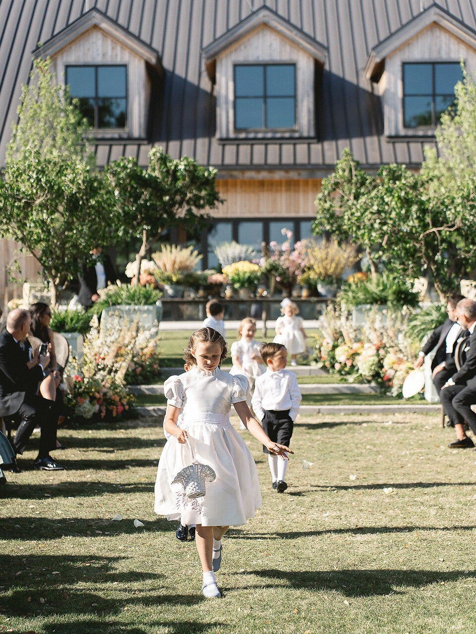 Children in formal attire page boy outfits and flower girl dresses walking outdoors with a building and garden in the background