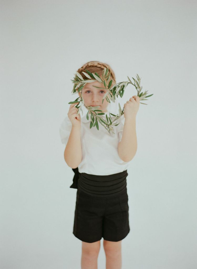 The Coco ensemble, a tuxedo-inspired outfit designed for stylish, modern flower girls. Features a double-lined white cotton shirt with a back row of pearl buttons, tailored tuxedo shorts, a satin cummerbund, and a French-wired bow. Interior waistline buttons secure the shirt, with a hand-tied sash for a polished finish. Handcrafted in Brooklyn from cotton and satin, with optional personalized embroidery. Perfect for weddings and special occasions.