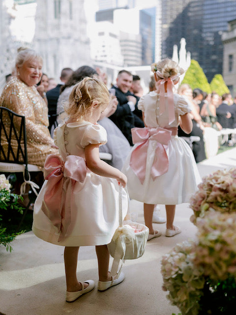 The Rachel VIP satin flower girl dress with pleated skirt, peter pan collar, and covered buttons – elegant choice for weddings and communions. With pop up sash