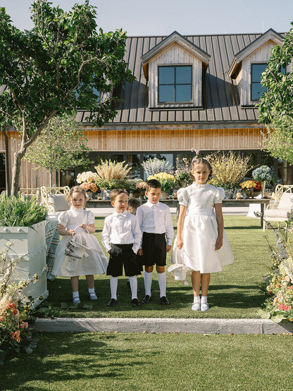 Children in formal attire page boy outfits and flower girl dresses standing in front of a house with decorative plants and furniture.