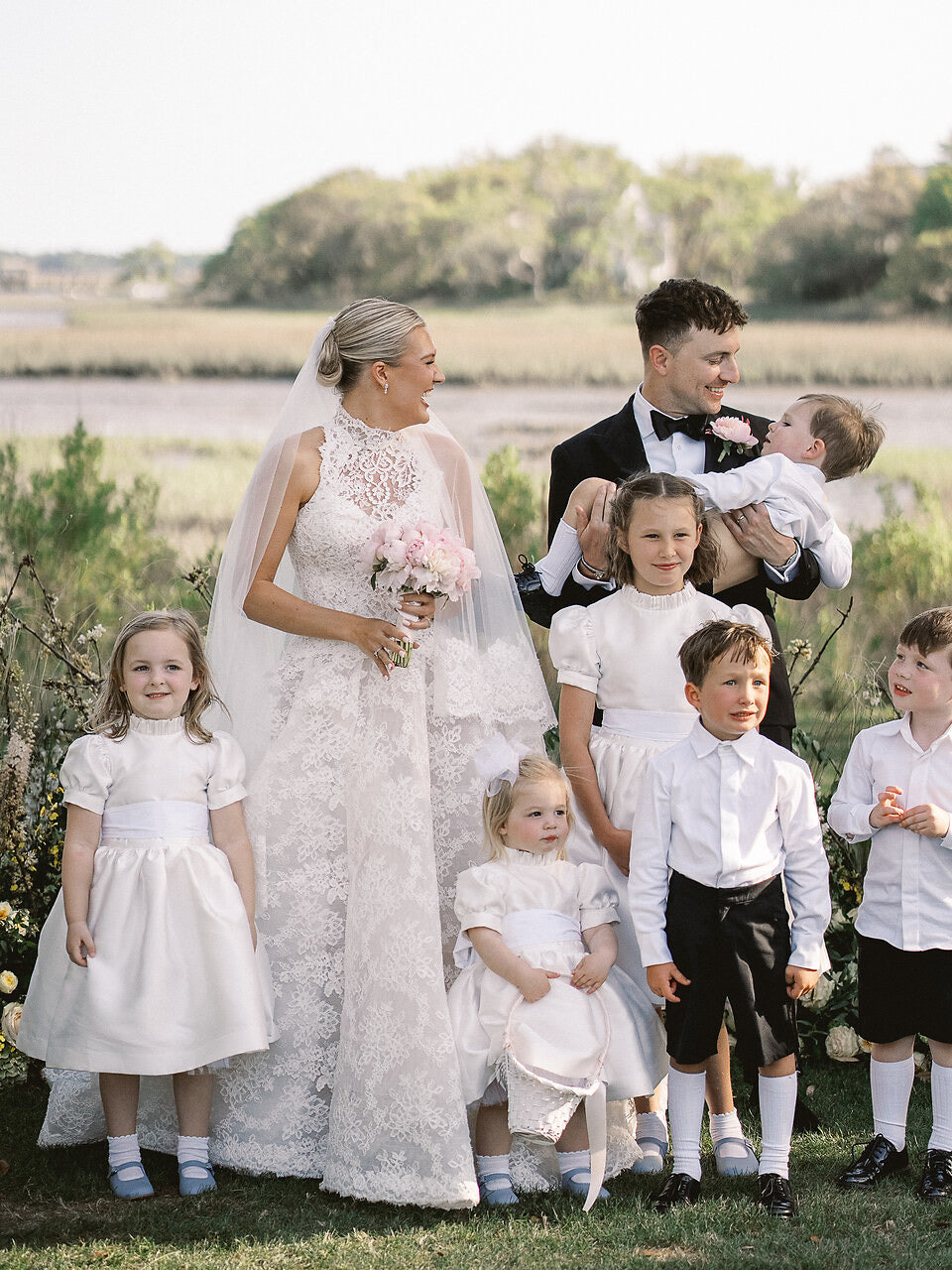 Wedding party with bride and groom holding a baby, surrounded by children in formal attire outdoors.