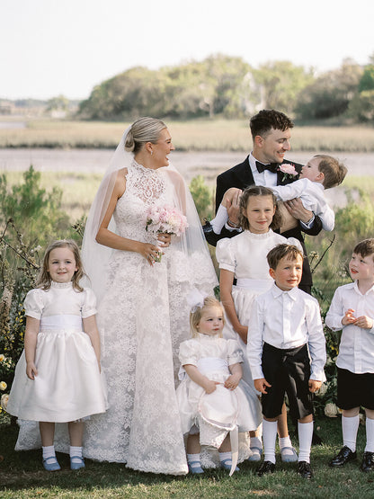 Wedding party with bride and groom holding a baby, surrounded by children in formal attire outdoors.