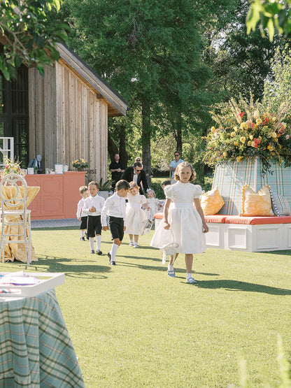 Children in white flower girl dresses and page boys outfits walking outdoors with a wooden structure and floral arrangement in the background.