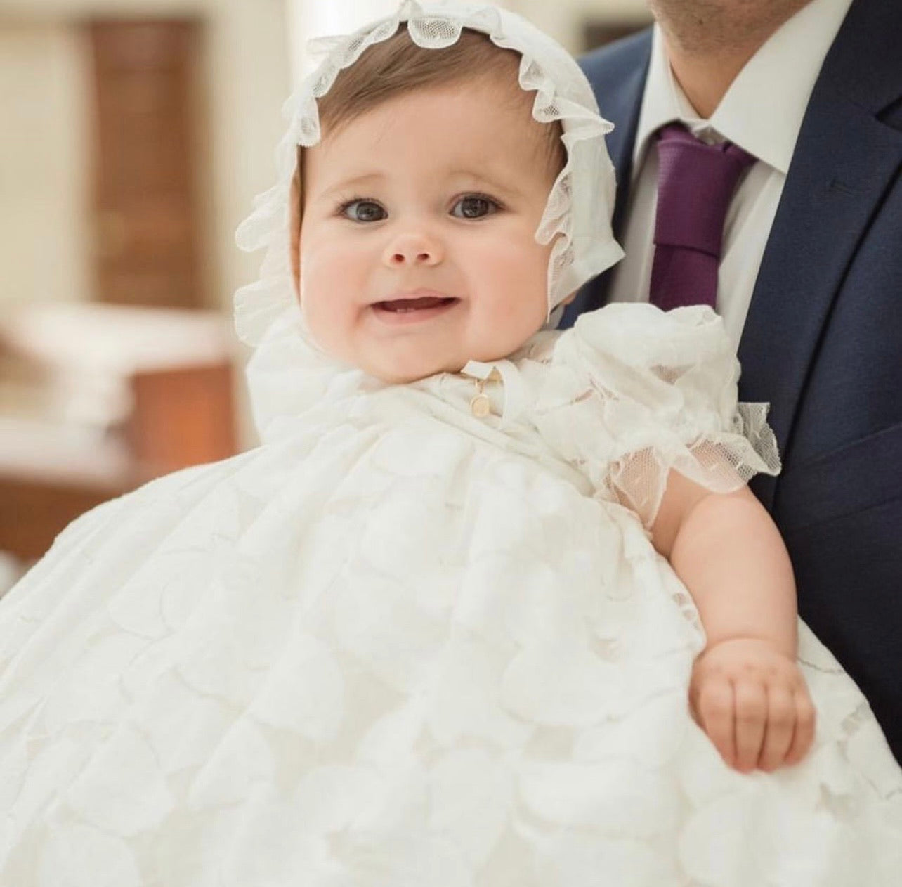 Infant wearing Doloris Petunia’s Baby Lace Ceremony Gown: hand‑cut floral cotton lace, point d’esprit edging, grosgrain bow at empire waist, button‑back closure, available in various skirt lengths, and paired with matching lace baby bonnet. Perfect for baptisms, christenings, and baby weddings.