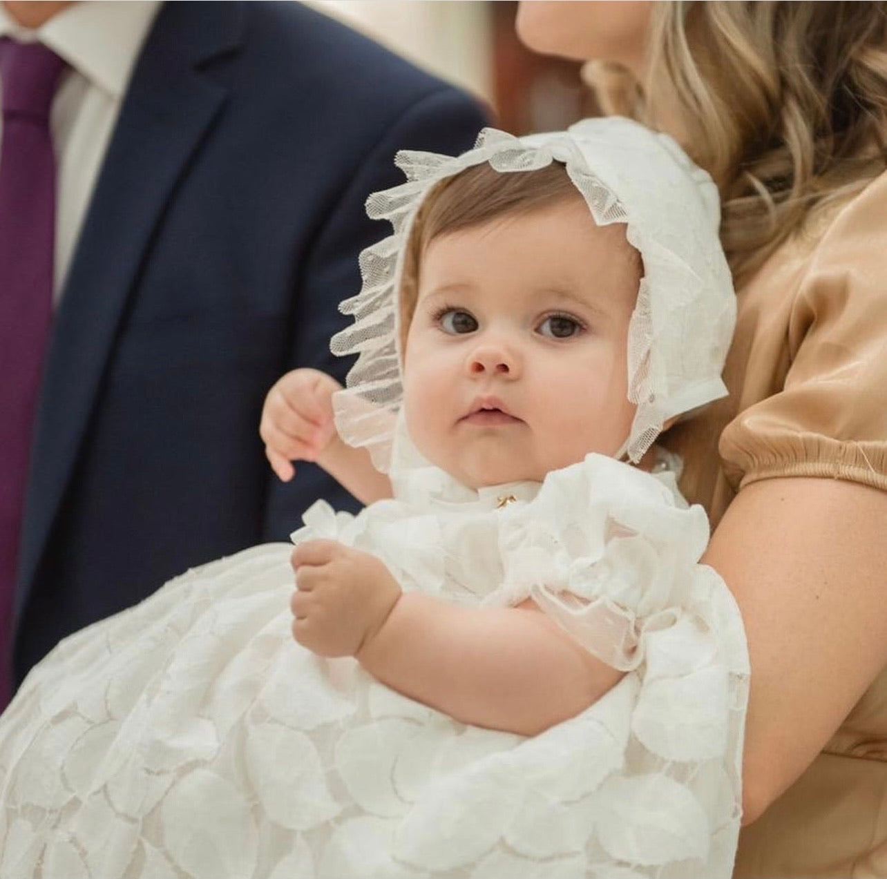 Infant wearing Doloris Petunia’s Baby Lace Ceremony Gown: hand‑cut floral cotton lace, point d’esprit edging, grosgrain bow at empire waist, button‑back closure, available in various skirt lengths, and paired with matching lace baby bonnet. Perfect for baptisms, christenings, and baby weddings.